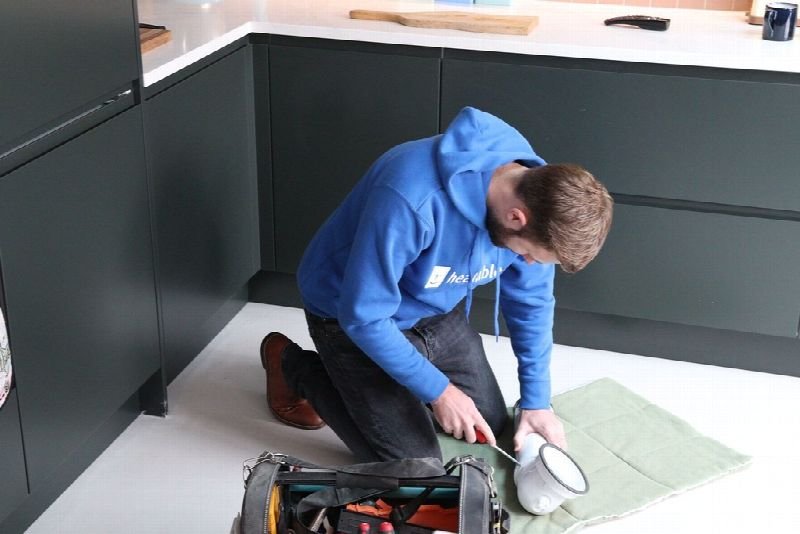 Tradesman in a blue jacket kneeling to work on a pipe fitting