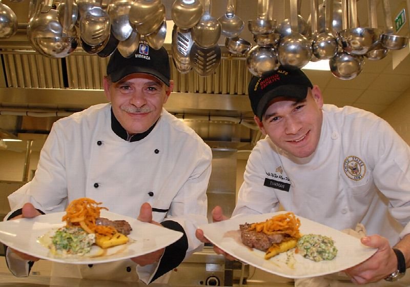 Two chefs in whites presenting plated dishes in a restaurant kitchen